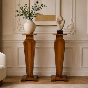 Op de afbeelding: Twee hoge, taps toelopende houten sokkels met vierkante voet en platte bovenkant. De linker sokkel draagt een witte keramische vaas met groen blad, de rechter sokkel draagt een lichtgekleurd abstract stenen beeld op een zwarte voet.