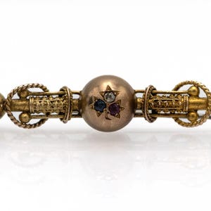 Może przedstawiać: Złota, zabytkowa broszka w kształcie paska z centralną kulą ozdobioną gwiazdką i trzema kamieniami szlachetnymi. Broszka ma skomplikowane detale, w tym akcenty w kształcie liny i ozdobne końcówki. Całkowity projekt jest symetryczny i bogato zdobiony.