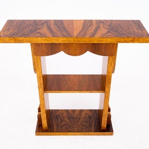 Puede incluir: Mesa auxiliar de madera con dos estantes y borde festoneado. La mesa tiene una tapa rectangular con un patrón de vetas de madera marrón oscuro. El marco y los estantes son de color marrón claro.