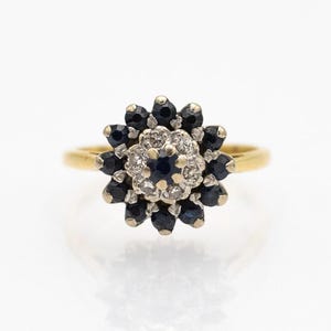 以下が含まれることがあります： 青いサファイアと白いダイヤモンドが花の形に集められた、金のリングです。