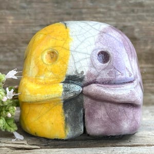 May include: A ceramic bird head sculpture, split into yellow and purple sections. The sculpture has a crackle glaze and a small eye hole. The beak is grey. Decorative art piece.