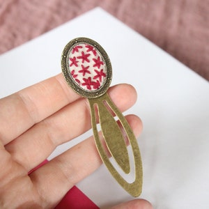 May include: A brass bookmark with a red embroidered floral design on a cream background. The bookmark has a decorative oval shape at the top and a long, thin, pointed tail.