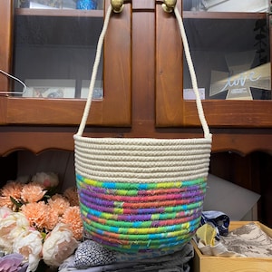 May include: A cream-coloured rope basket with a colourful, woven design. The basket has a rainbow pattern of pink, green, purple, and blue. It has a cream-coloured rope handle and is hanging from a wooden cabinet.