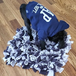 May include: A black dog wearing a navy blue GAP sweatshirt with white lettering, standing on a grey and white fabric mat. The mat is made of strips of fabric in varying shades of grey and white.