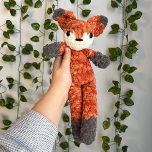 May include: A handmade crochet fox doll with orange, white, and gray yarn. The fox has a fluffy texture, with gray accents on the ears, paws, and feet. The doll is held up against a background of green leaves.