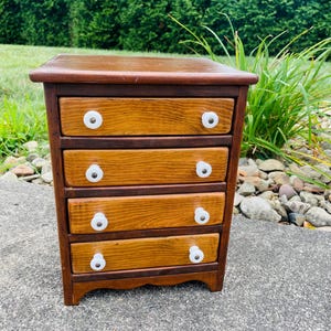 May include: A small, wooden chest of drawers with four drawers. The chest has a dark brown frame and a lighter wood front. Each drawer has a white, round knob. The chest is outdoors, in front of green foliage.