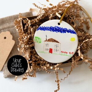 May include: A white ceramic ornament featuring a child's drawing of a house with a red door, blue sky, and yellow sun. The ornament is in a box with brown paper filler, a burlap bag, and a tag. The text "Your Child's Drawing" is on a black circle.