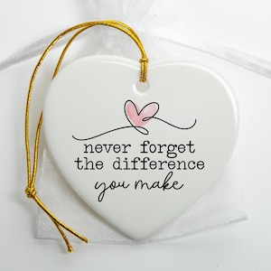 May include: A white heart-shaped ceramic ornament featuring a small pink heart illustration and the black text "never forget the difference you make". The ornament includes a gold hanging cord and is placed on a sheer white fabric pouch.