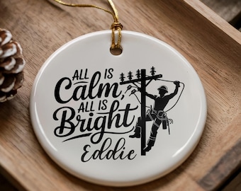 Może przedstawiać: Okrągła biała ceramiczna ozdoba z czarnym napisem All is Calm, All is Bright oraz imieniem Eddie. Wzór zawiera czarną sylwetkę pracownika energetyki wspinającego się na słup elektryczny z przewodami i oprzyrządowaniem.