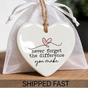 May include: A white heart-shaped ceramic ornament hanging from a brown twine string in front of a white organza gift bag. The ornament features a small pink heart graphic and the black text: never forget the difference you make.
