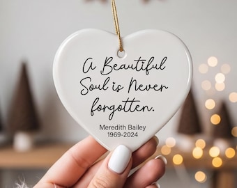 May include: A white heart-shaped ceramic ornament featuring the black printed text A Beautiful Soul is Never forgotten. Below the text, the name Meredith Bailey and the years 1969-2024 are printed. The ornament hangs from a thin gold string.