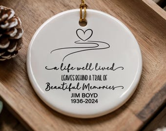 May include: A round white ceramic ornament featuring a black line drawing of a heart and a winding path. The text reads: A life well lived leaves behind a trail of beautiful memories, followed by the name JIM BOYD and the years 1936-2024.