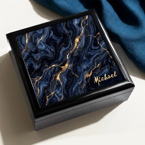 May include: A square black wooden box featuring a lid with a dark blue and black marble pattern accented by gold veins. The name Michael is printed in gold script in the bottom right corner of the lid, which has a smooth, polished finish.