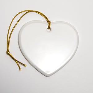May include: A smooth, white, heart-shaped ceramic ornament with a glossy finish. The ornament features a small hole at the top center, through which a thin, metallic gold cord is looped and knotted for hanging purposes.
