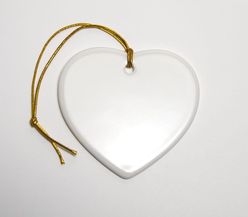 May include: A smooth, white, heart-shaped ceramic ornament with a glossy finish. The ornament features a small hole at the top center, through which a thin, metallic gold cord is looped and knotted for hanging purposes.
