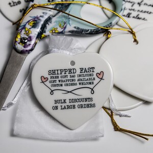 May include: A white ceramic heart-shaped ornament with black text reading SHIPPED FAST, FREE GIFT BAG INCLUDED, GIFT WRAPPING AVAILABLE, CUSTOM ORDERS WELCOME, BULK DISCOUNTS ON LARGE ORDERS, and two small pink hearts, next to floral scissors.