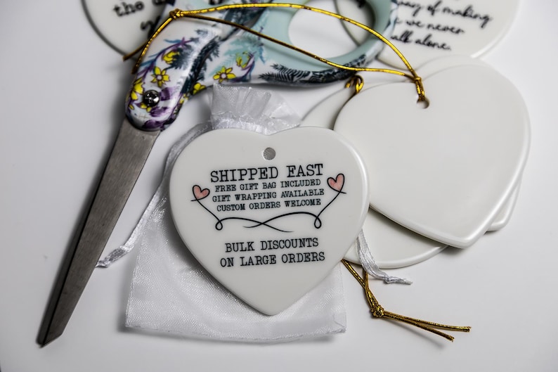 May include: A white ceramic heart-shaped ornament with black text reading SHIPPED FAST, FREE GIFT BAG INCLUDED, GIFT WRAPPING AVAILABLE, CUSTOM ORDERS WELCOME, BULK DISCOUNTS ON LARGE ORDERS, and two small pink hearts, next to floral scissors.