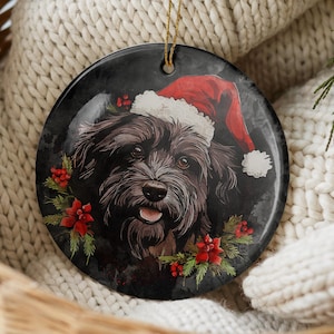 May include: A round ceramic ornament featuring a black dog wearing a red and white Santa hat. The dog is surrounded by green pine needles and red holly berries against a dark, smoky background. A gold string is attached to the top for hanging.