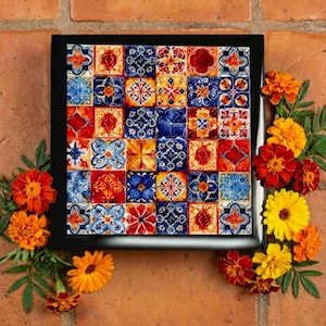 May include: A framed square tile art piece with a black frame. The tiles are a mix of red, blue, yellow, and white, with intricate floral and geometric patterns. Orange and yellow marigold flowers and green leaves surround the frame.