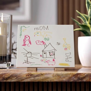 May include: A white ceramic tile with colorful childlike drawings and the words "MOM" and "Dad". The drawings include a house, birds, and other whimsical figures. The tile is displayed on a wooden stand.
