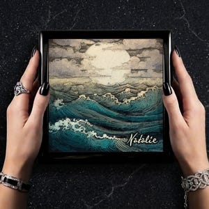 May include: A framed artwork depicting a seascape with large waves under a cloudy sky and a full moon. The artwork is signed "Natalie" in cursive. The frame is black.