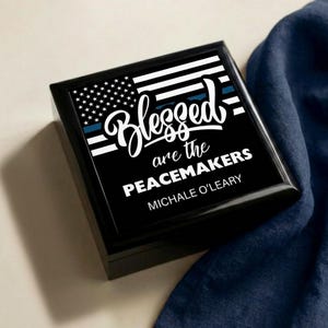 May include: Black square box with a glossy finish. The top features a design of the American flag with a blue line, the words "Blessed are the Peacemakers," and the name "Michale O'Leary" in white script and block letters.