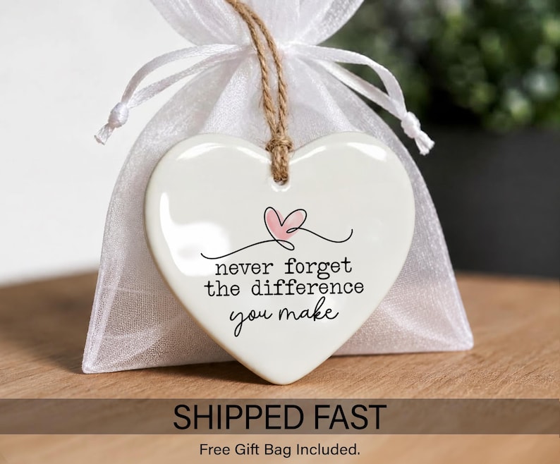 May include: A white heart-shaped ceramic ornament hanging from a brown twine string in front of a white organza gift bag. The ornament features a small pink heart graphic and the black text: never forget the difference you make.