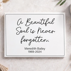 May include: A white rectangular sign with a black border features the text "A Beautiful Soul is Never Forgotten." Below the quote is "Meredith Bailey 1969-2024" in black script. The sign is surrounded by decorative elements.