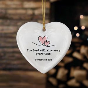 May include: A white heart-shaped ceramic ornament hanging from a gold string. The center features two small pink hearts outlined in black, with the text: The Lord will wipe away every tear. Revelation 21:4 printed in black lettering below the hearts.