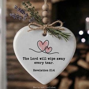 May include: White heart-shaped ceramic ornament with the text "The Lord will wipe away every tear. Revelation 21:4." Features a pink heart design and is adorned with dried herbs and twine. Includes a wooden bead hanger.