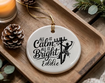 以下が含まれることがあります： 白いセラミック製のクリスマスオーナメント。黒い筆記体で「All is Calm, All is Bright, Eddie」と書かれています。電柱で作業する電気技師のシルエットも特徴です。オーナメントは金の紐から吊り下げられています。