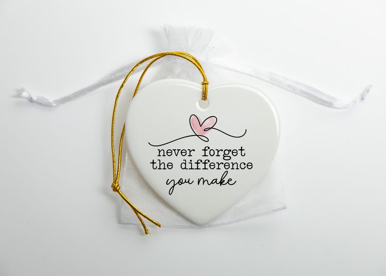 May include: A white heart-shaped ceramic ornament featuring a small pink heart illustration and the black text "never forget the difference you make". The ornament includes a gold hanging cord and is placed on a sheer white fabric pouch.
