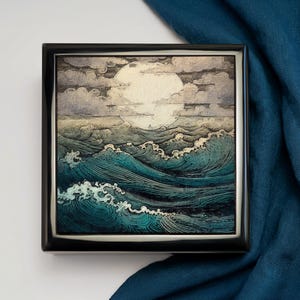 May include: A square, black jewelry box with a detailed illustration of ocean waves under a full moon. The artwork features shades of teal, gray, and white, creating a serene seascape scene. The box is displayed on a blue fabric.