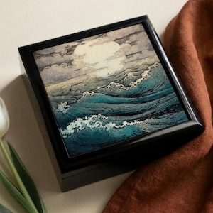 May include: A black wooden box with a framed image of ocean waves under a full moon. The artwork features shades of blue, teal, and white, with a large, bright moon in the sky. The box is rectangular and sits on a light-colored surface.