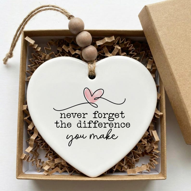 May include: A white heart-shaped ceramic ornament featuring a small pink heart illustration and the black text "never forget the difference you make." The ornament hangs from a jute twine string with three wooden beads.