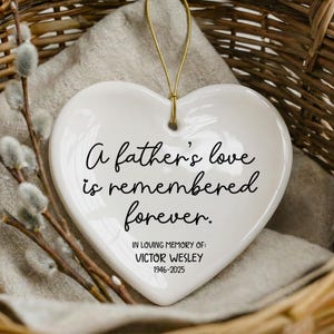 May include: White heart-shaped ceramic ornament with gold string, inscribed with "A father's love is remembered forever." Includes "In Loving Memory of: VICTOR WESLEY 1946-2025." Resting in a woven basket with sprigs of pussy willow.