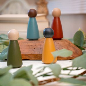 Puede incluir: Cinco figuritas de madera con cuerpos y cabezas de diferentes colores. Las figuritas están de pie sobre una superficie de madera. Las figuritas están rodeadas de hojas verdes.