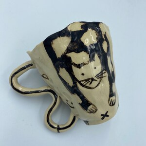 Könnte beinhalten: Eine Keramiktasse mit einem schwarz-weißen Katzendesign. Die Tasse hat ein einzigartiges, handgefertigtes Aussehen und verfügt über einen Doppelgriff.