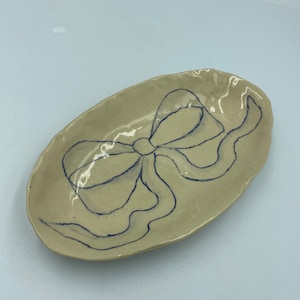 May include: A white ceramic oval dish with a blue hand-drawn bow design.