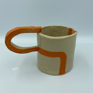 Op de afbeelding: Een keramische mok met een crèmekleurige glazuur en een oranje handvat. De mok heeft een geometrisch ontwerp met oranje lijnen.
