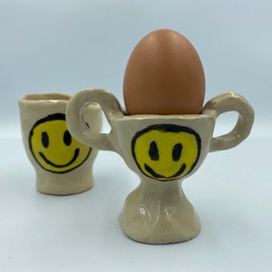 Könnte beinhalten: Zwei Keramik-Eierbecher mit einem Smiley-Gesichtsdesign. Ein Eierbecher hält ein braunes Ei.