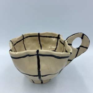 Könnte beinhalten: Eine weiße Keramiktasse mit einem schwarzen Gittermuster und einem Henkel. Die Tasse hat eine leicht unregelmäßige Form.