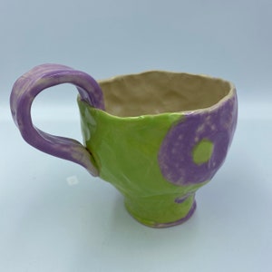 Könnte beinhalten: Eine handgefertigte Keramiktasse mit einem grün-violetten, gesprenkelten Überzug. Die Tasse hat eine einzigartige, asymmetrische Form und einen großen Henkel.