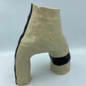 Könnte beinhalten: Eine Keramikvase mit einer einzigartigen, abstrakten Form. Die Vase ist cremefarben mit schwarzen, bemalten Linien. Die Vase hat eine breite Öffnung und einen schmalen Boden.