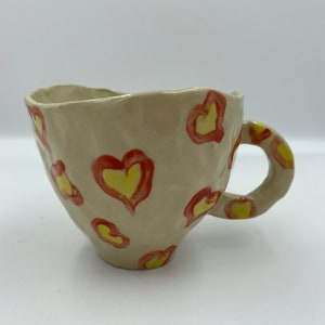 May include: A handmade ceramic mug with a textured surface and a handle. The mug is decorated with red and yellow hearts.