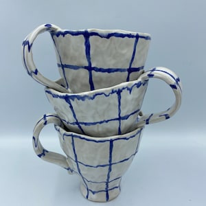Könnte beinhalten: Drei weiße Keramikbecher mit blauen Gittermustern, die übereinander gestapelt sind. Die Becher haben eine leicht strukturierte Oberfläche und eine abgerundete Form.