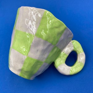 Peut inclure: Une tasse en céramique avec un motif à carreaux dans des tons de vert et de violet. La tasse a une surface unique, inégale et texturée.