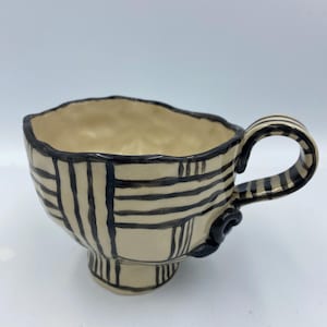 Könnte beinhalten: Eine handgefertigte Keramiktasse mit einem geometrischen Muster in Schwarz und Weiß. Die Tasse hat eine einzigartige, leicht unregelmäßige Form und einen Griff mit abgerundetem Ende.