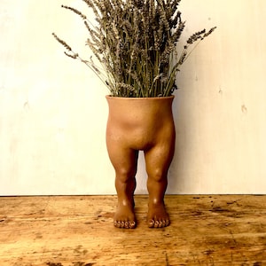Puede incluir: Un macetero de terracota con forma de piernas con flores de lavanda secas en su interior.