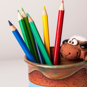 以下が含まれることがあります： 白い顔と茶色の体をした羊の形をしたセラミック製の鉛筆立て。鉛筆立ては青と茶色のストライプのベースがあり、赤、黄、緑、青など、色鉛筆が入っています。